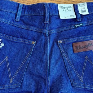May include: Close-up of the back of blue Wrangler Retro Bailey high-rise trousers. The jeans have embroidered floral details on the pockets and a brown leather Wrangler patch. The Wrangler label is visible.