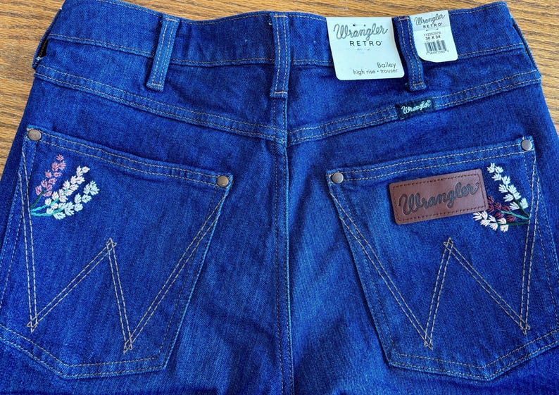 May include: Close-up of the back of blue Wrangler Retro Bailey high-rise trousers. The jeans have embroidered floral details on the pockets and a brown leather Wrangler patch. The Wrangler label is visible.