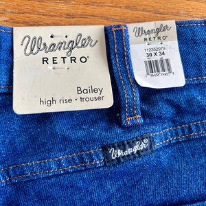May include: Close-up of blue Wrangler Retro Bailey high-rise trousers. The jeans have a dark blue wash with orange stitching. A tag reads "Wrangler RETRO Bailey high rise &bull; trouser" and another tag shows the size 30 x 34.