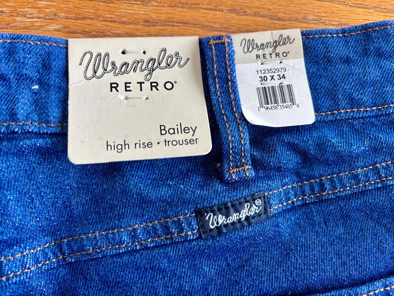 May include: Close-up of blue Wrangler Retro Bailey high-rise trousers. The jeans have a dark blue wash with orange stitching. A tag reads "Wrangler RETRO Bailey high rise &bull; trouser" and another tag shows the size 30 x 34.