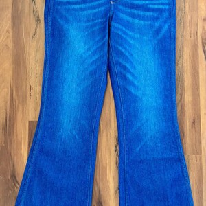 May include: Blue denim jeans with a high waist and flared legs. The jeans have a classic five-pocket design and are embellished with small pink floral embroidery near the front pockets. The denim has a faded wash effect.