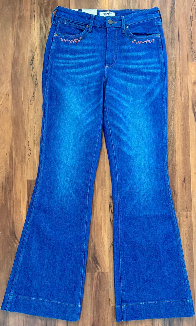 May include: Blue denim jeans with a high waist and flared legs. The jeans have a classic five-pocket design and are embellished with small pink floral embroidery near the front pockets. The denim has a faded wash effect.