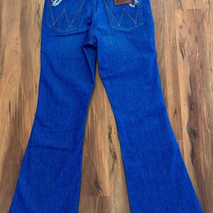 May include: Blue denim Wrangler jeans with a flared leg. The back pockets have a floral embroidery design. The Wrangler logo is on a brown leather patch. The jeans have a high waist and a classic five-pocket design.