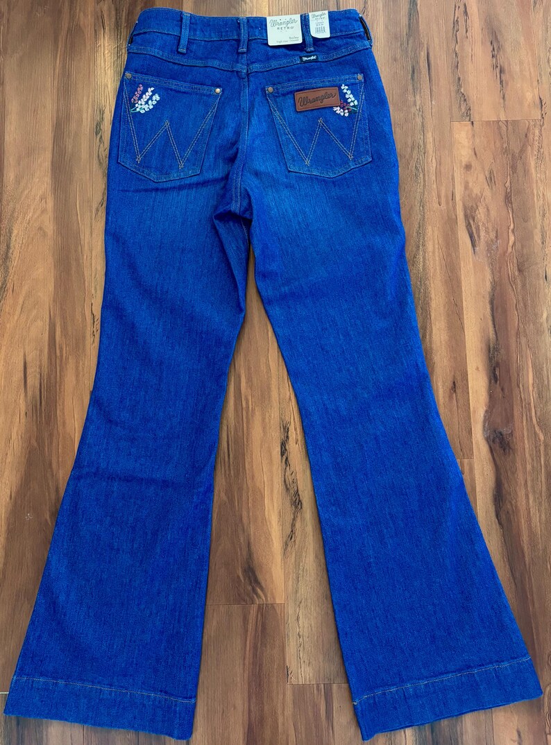 May include: Blue denim Wrangler jeans with a flared leg. The back pockets have a floral embroidery design. The Wrangler logo is on a brown leather patch. The jeans have a high waist and a classic five-pocket design.