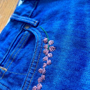 May include: Close-up of blue denim jeans with a floral embroidery design. The embroidery features pink flowers and green stems. The jeans have a classic five-pocket design with metal rivets and a button closure.