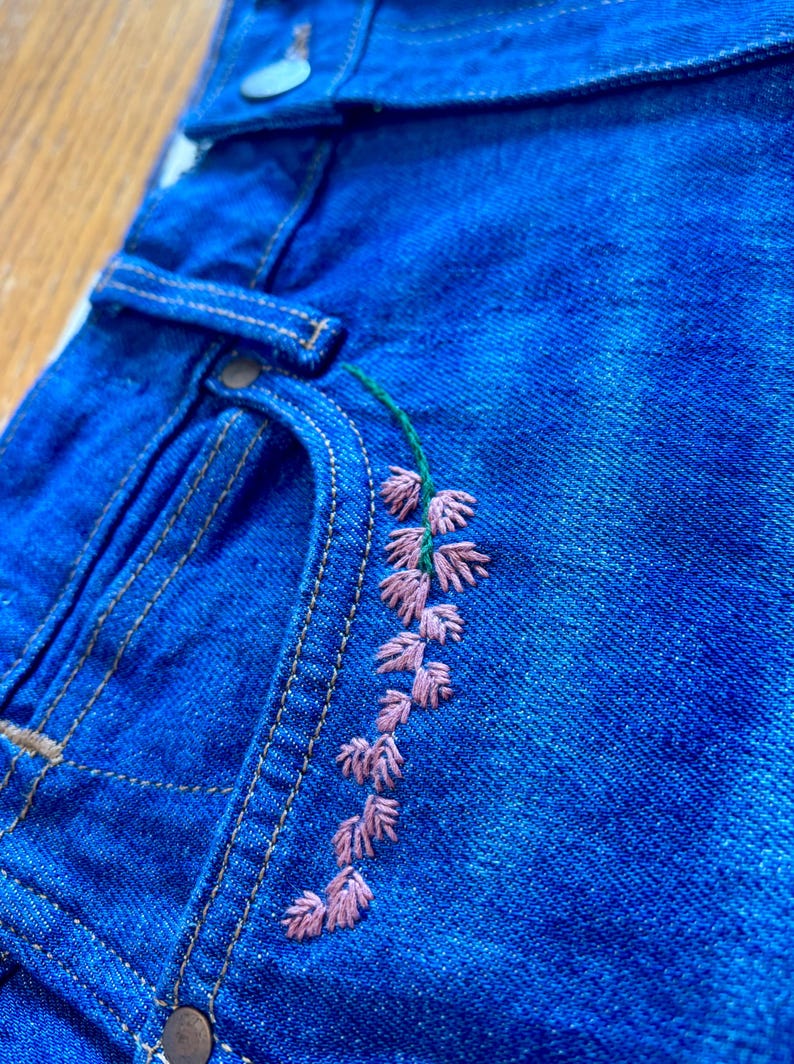 May include: Close-up of blue denim jeans with a floral embroidery design. The embroidery features pink flowers and green stems. The jeans have a classic five-pocket design with metal rivets and a button closure.