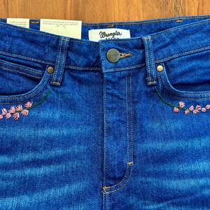 May include: Close-up of blue denim jeans with pink floral embroidery on the front pockets. The jeans have a button closure and the brand "Wrangler" is visible on the waistband.