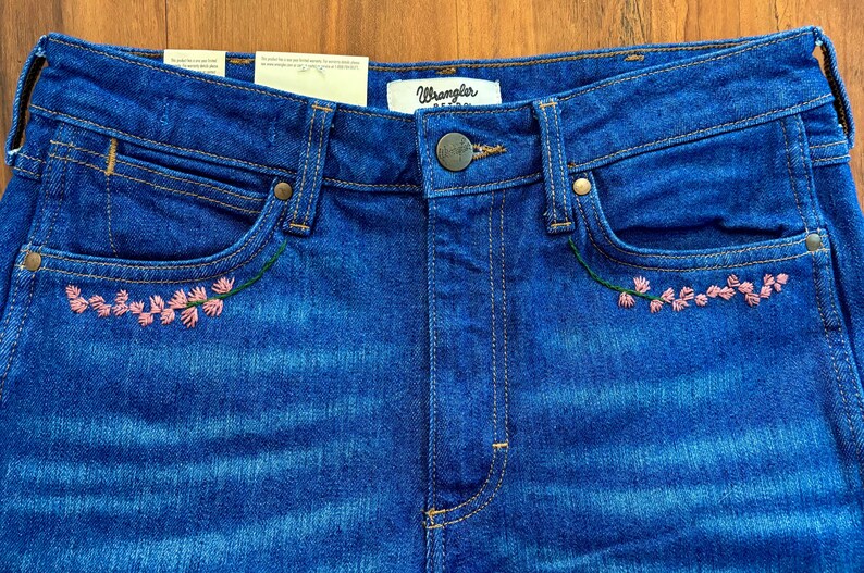 May include: Close-up of blue denim jeans with pink floral embroidery on the front pockets. The jeans have a button closure and the brand "Wrangler" is visible on the waistband.