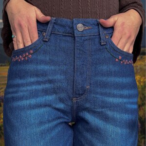 May include: Close-up of blue denim jeans with floral embroidery on the front pockets. The jeans have a button closure and classic five-pocket design. The pockets are accented with pink floral embroidery and green stitching.