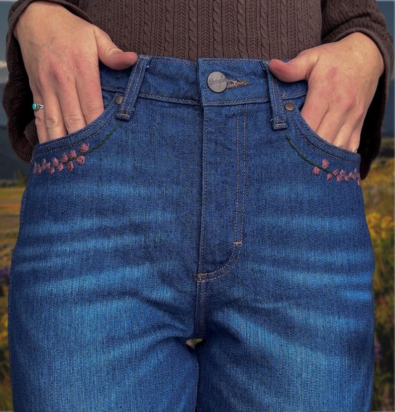 May include: Close-up of blue denim jeans with floral embroidery on the front pockets. The jeans have a button closure and classic five-pocket design. The pockets are accented with pink floral embroidery and green stitching.