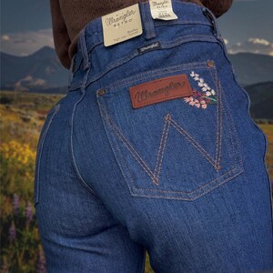 May include: Blue Wrangler Retro Bailey high-rise jeans with a floral embroidered design on the back pocket. The jeans feature a brown leather patch with the Wrangler logo and a classic five-pocket design.