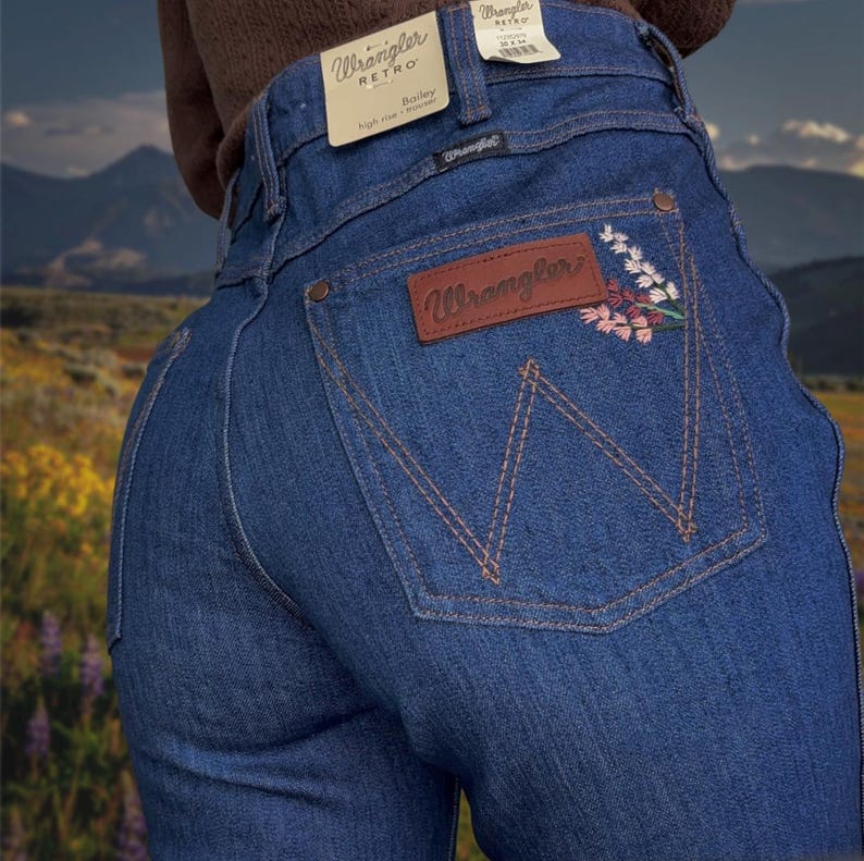 May include: Blue Wrangler Retro Bailey high-rise jeans with a floral embroidered design on the back pocket. The jeans feature a brown leather patch with the Wrangler logo and a classic five-pocket design.