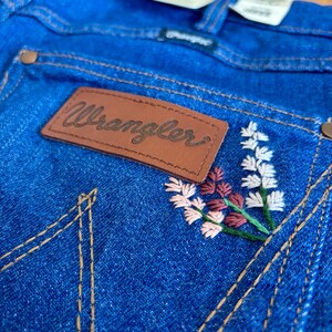 May include: Close-up of blue denim jeans with a brown leather patch that reads "Wrangler." The jeans feature embroidered floral details in white, pink, and burgundy. The stitching is in a contrasting tan color.