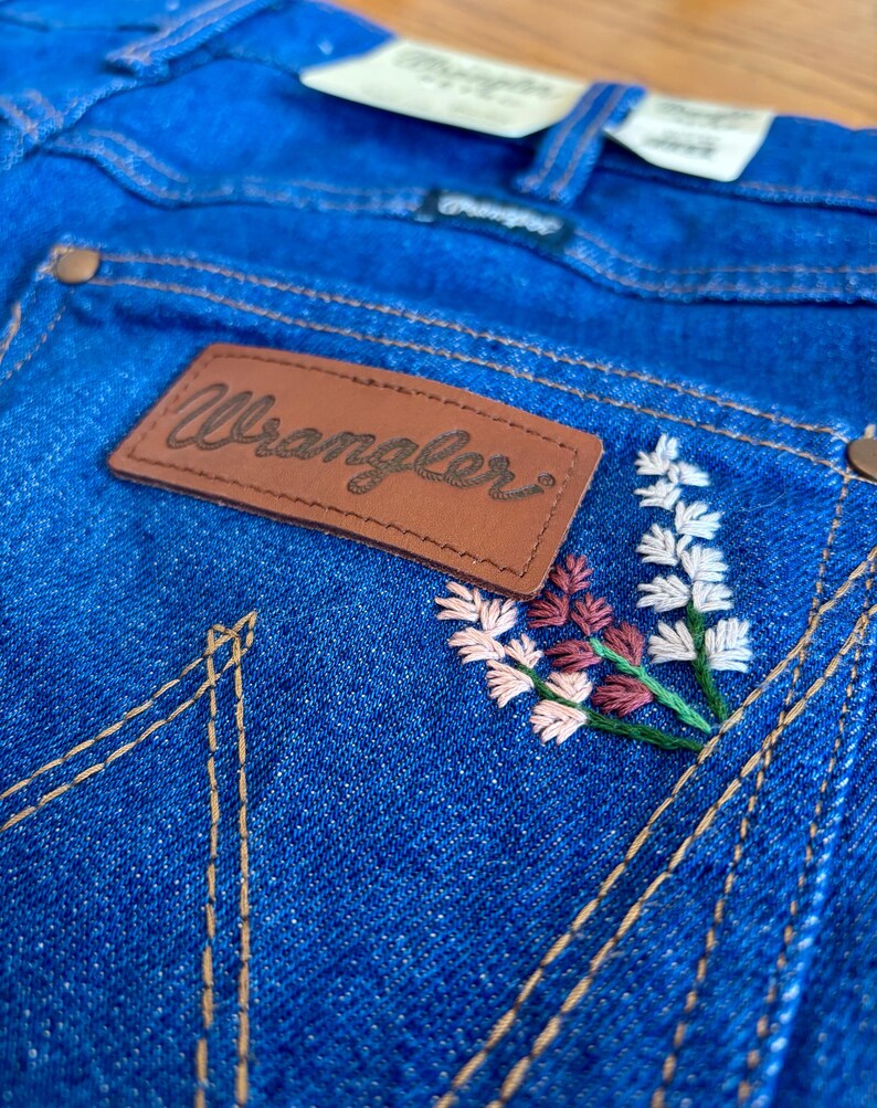 May include: Close-up of blue denim jeans with a brown leather patch that reads "Wrangler." The jeans feature embroidered floral details in white, pink, and burgundy. The stitching is in a contrasting tan color.