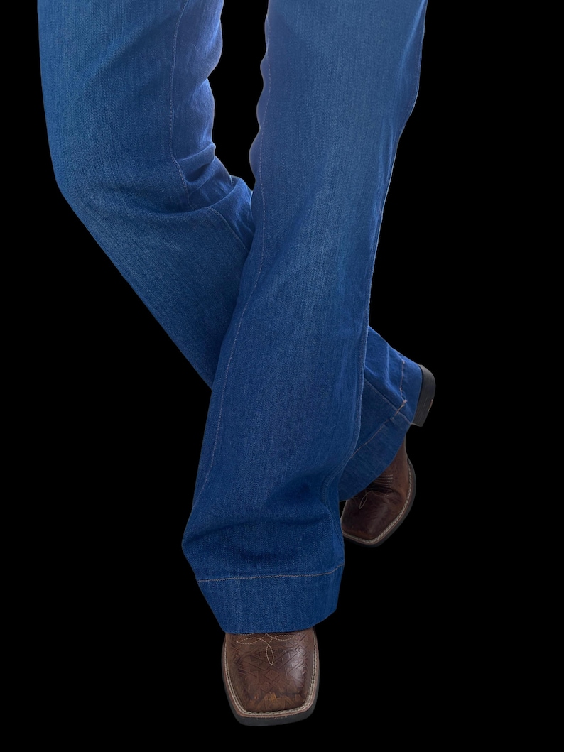 May include: Blue denim jeans with a flared leg design. The jeans are paired with brown leather square-toe boots. The image is set against a black background, highlighting the clothing and footwear.