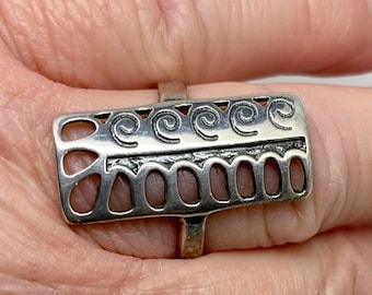 May include: A silver ring with a rectangular design featuring intricate swirls and cutouts. The ring is worn on a finger.