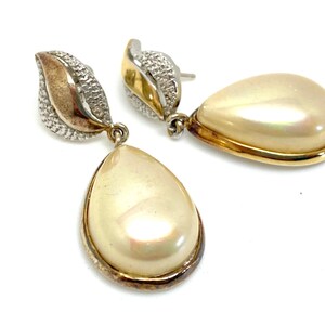 Puede incluir: Un par de pendientes colgantes de tono dorado y plateado con piedras en forma de lágrima nacaradas.