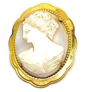 Puede incluir: Un broche de camafeo dorado con una talla de camafeo blanco de un perfil de mujer. El camafeo está engastado en un marco ovalado dorado con un borde de cuerda.