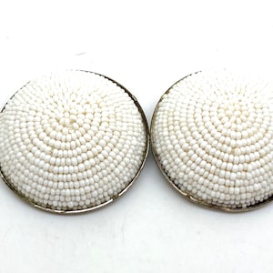 May include: A pair of white beaded earrings with a silver rim. The earrings are round and have a dome shape.