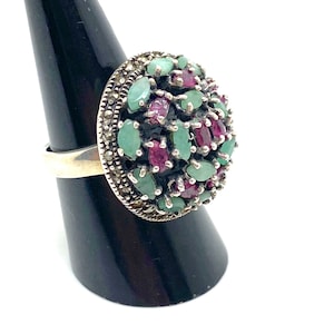 May include: A silver ring with a large, round cluster of green and red gemstones. The ring is set on a black ring holder.