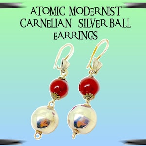Pode incluir: Um par de brincos de bola de prata com pedras de cornalina. Os brincos são suspensos por um gancho de prata e têm um berloque de bola de prata abaixo da pedra de cornalina. Os brincos são chamados de "Atomic Modernist Carnelian Silver Ball Earrings".