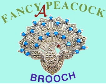 May include: A silver peacock brooch with blue and red stones. The brooch is in the shape of a peacock with its tail feathers spread out. The text "FANCY A PEACOCK BROOCH" is written around the brooch.