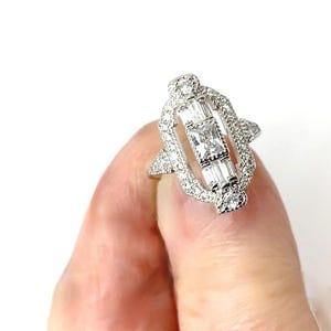 Pode incluir: Um anel de prata com um design retangular, apresentando uma pedra central quadrada e pedras de corte baguette. O anel &eacute; incrustado com pequenas pedras redondas, criando um efeito brilhante. O anel &eacute; segurado por um dedo.
