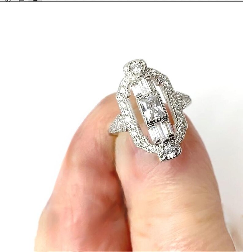 Pode incluir: Um anel de prata com um design retangular, apresentando uma pedra central quadrada e pedras de corte baguette. O anel &eacute; incrustado com pequenas pedras redondas, criando um efeito brilhante. O anel &eacute; segurado por um dedo.