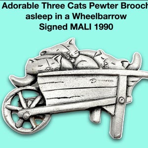 May include: A silver pewter brooch depicting three cats asleep in a wheelbarrow. The brooch is signed "MALI 1990".