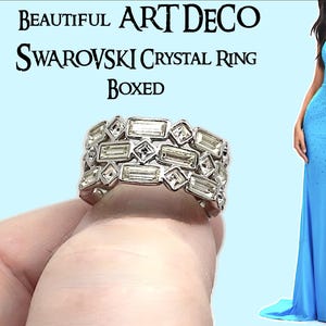May include: A silver-toned Art Deco ring with multiple rectangular and square crystals. The ring is held up by a hand. The text "BEAUTIFUL ART DECO SWAROVSKI CRYSTAL RING BOXED" is displayed above the ring. A woman in a blue dress is in the background.