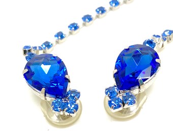 May include: A pair of blue teardrop-shaped earrings with smaller blue rhinestones surrounding the larger stones. The earrings are attached to silver posts.