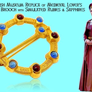 Puede incluir: Broche dorada con rubíes y zafiros simulados. La broche circular tiene un cierre de alfiler y está adornada con piedras preciosas rojas y azules alternas. El texto dice "British Museum Replica of Medieval Lover's Gold Brooch".