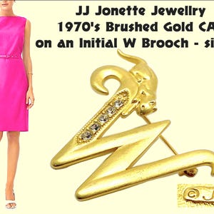 May include: A gold-toned brooch featuring a stylised letter 'W' with a brushed gold finish. A small cat figurine is perched on top of the letter, and the brooch is adorned with clear rhinestones. The brooch is signed 'JJ' on the back.