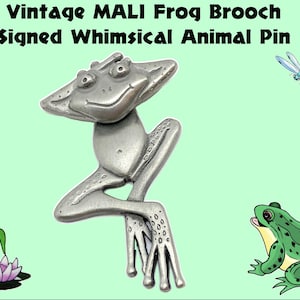 Op de afbeelding: Een vintage zilverkleurige kikkerbroche met de tekst "Vintage MALI Frog Brooch Signed Whimsical Animal Pin". De kikkerpin heeft een grillig ontwerp en staat tegen een lichtgroene achtergrond met illustraties van een libelle en een kikker.