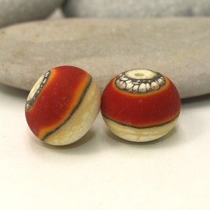 May include: Two round beads with a cream-colored base and a vibrant red band. One bead has a decorative pattern in black and white. The beads are likely for jewelry making or crafts.