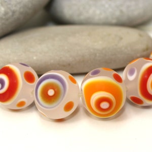 Puede incluir: Primer plano de varias cuentas redondas de vidrio con una base de color crema. Las cuentas presentan círculos concéntricos en naranja, rojo y morado, con puntos más pequeños en naranja y morado. Las cuentas están junto a piedras grises lisas.