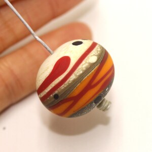 May include: A round, multi-coloured glass bead with a white, red, orange and brown striped pattern. The bead has black dots and is strung on a wire.