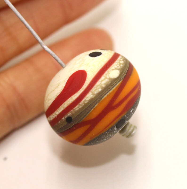 May include: A round, multi-coloured glass bead with a white, red, orange and brown striped pattern. The bead has black dots and is strung on a wire.