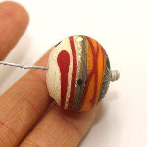 May include: A round, multi-coloured glass bead with a white, red, orange, and brown swirl pattern. The bead has a small hole in the side for stringing.