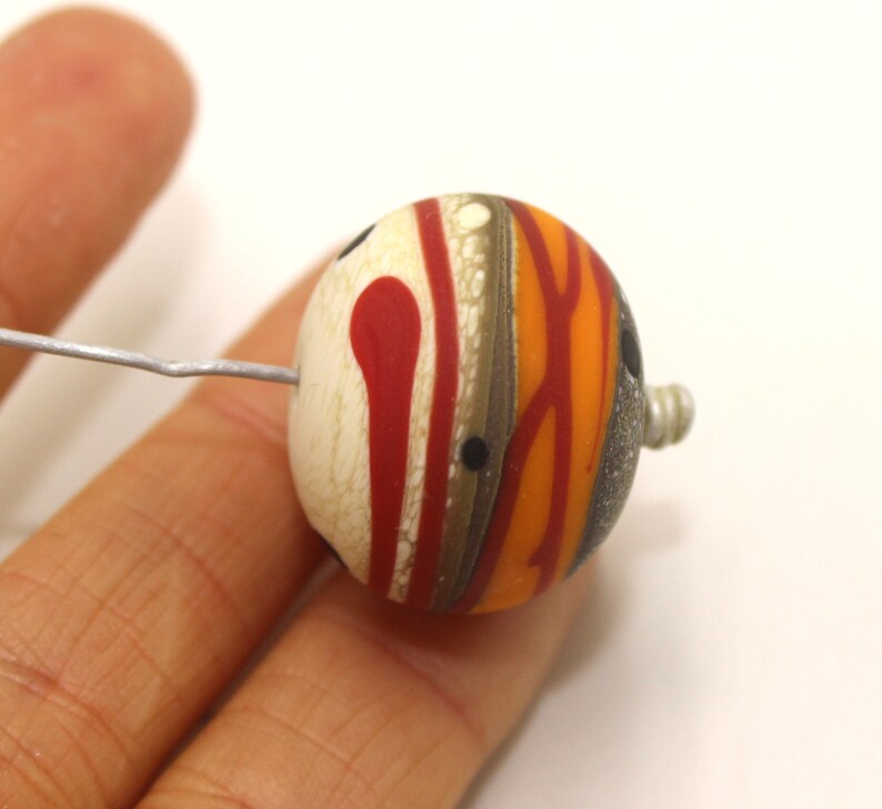 May include: A round, multi-coloured glass bead with a white, red, orange, and brown swirl pattern. The bead has a small hole in the side for stringing.