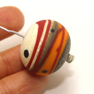May include: A round, multi-coloured glass bead with a white, red, orange, brown, and grey swirl pattern. The bead has black dots on the surface and a silver wire loop attached to the side.