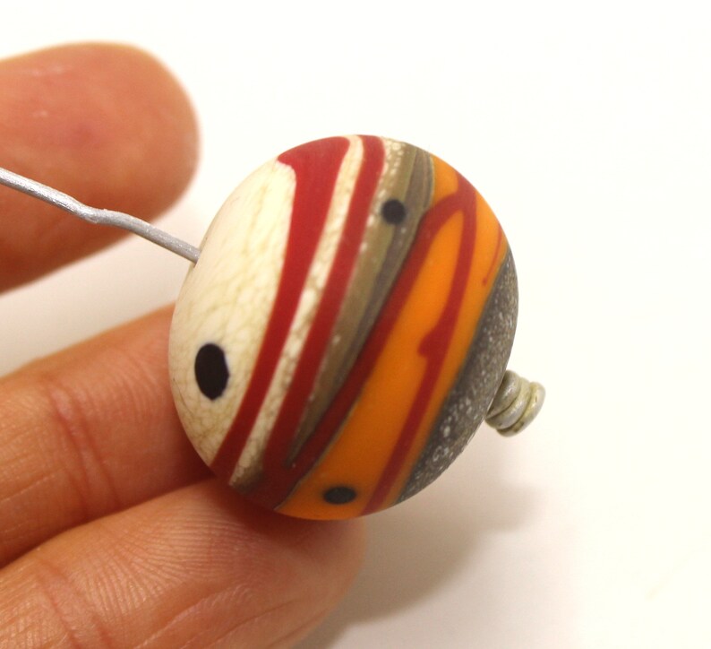 May include: A round, multi-coloured glass bead with a white, red, orange, brown, and grey swirl pattern. The bead has black dots on the surface and a silver wire loop attached to the side.