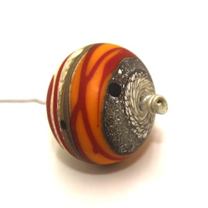 May include: A round, orange, red, white and brown glass bead with a silver metal loop. The bead has a swirl pattern and a hole in the centre.