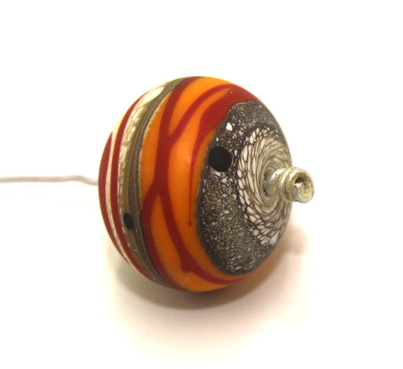 May include: A round, orange, red, white and brown glass bead with a silver metal loop. The bead has a swirl pattern and a hole in the centre.