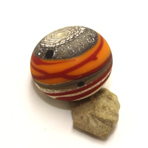 May include: A round glass bead with a swirling pattern of orange, red, brown, and white. The bead has a small hole in the centre.
