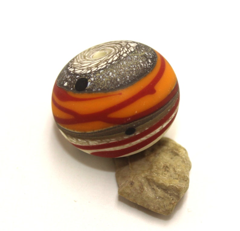 May include: A round glass bead with a swirling pattern of orange, red, brown, and white. The bead has a small hole in the centre.
