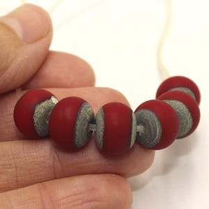 Könnte beinhalten: Eine Kette aus roten Perlen mit mattem Finish, jede mit einem grau-weißen Akzent. Die runden Perlen sind auf einer dünnen, cremefarbenen Schnur aufgefädelt. Die Kette wird in einer Hand gehalten.