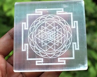 Puede incluir: Un cubo de cristal transparente con un símbolo Sri Yantra grabado en blanco. El símbolo es un patrón geométrico de triángulos y cuadrados.