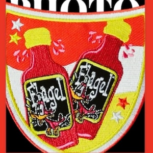 Könnte beinhalten: Ein gestickter Patch mit zwei roten Flaschen scharfer Sauce. Die Flaschen haben ein schwarzes Etikett mit dem Wort "Fligel" darauf gedruckt. Der Patch ist schildförmig und hat einen weißen Rand.