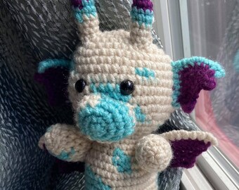 May include: A handmade crochet dragon plushie in cream, teal, and purple. The dragon has large black eyes, small wings, and horns. The plushie is sitting in front of a window.